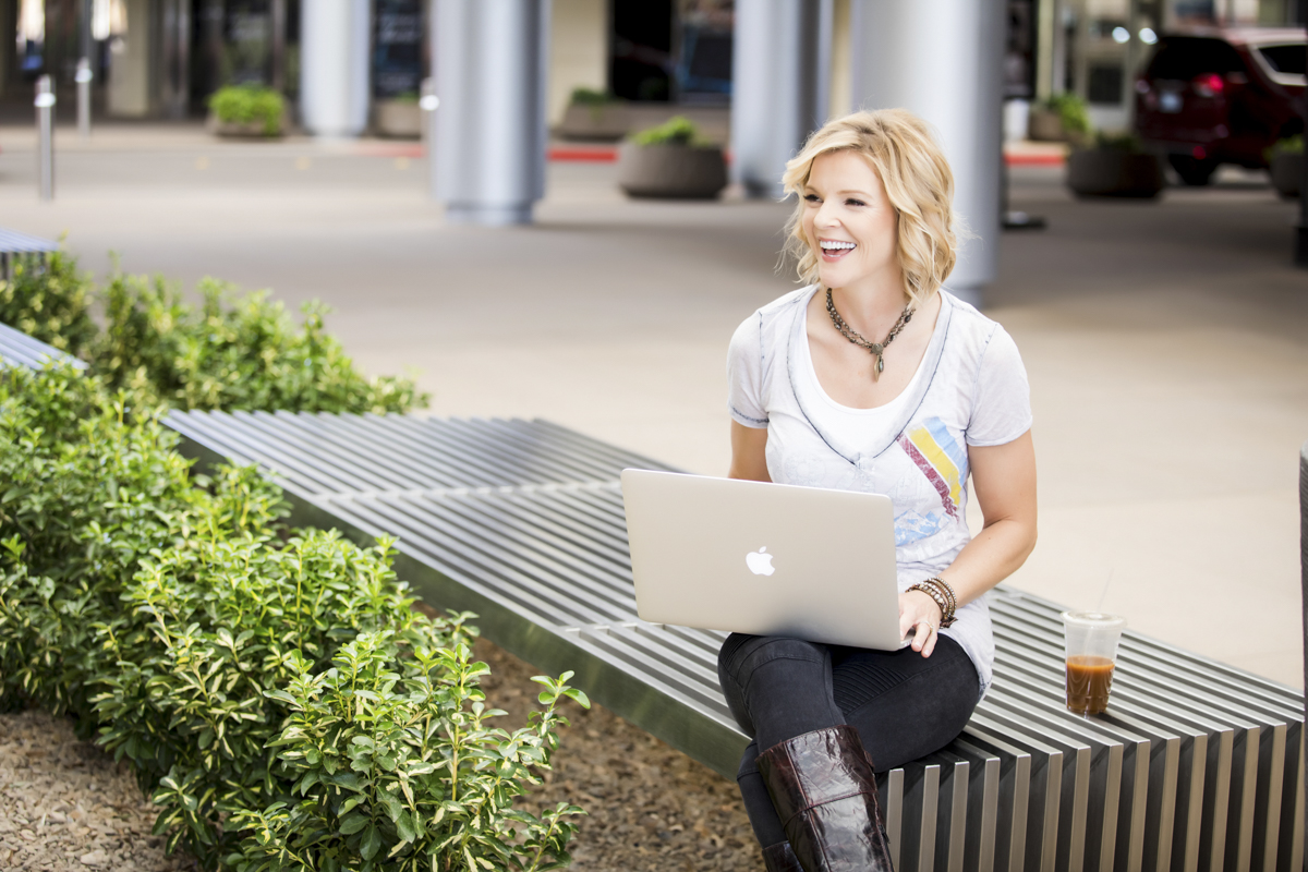 Woman sitting on bench smiling with laptop open and drink next to her.
