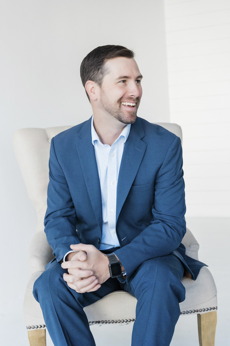 Man in blue suit sitting down and smiling with hands together over lap.